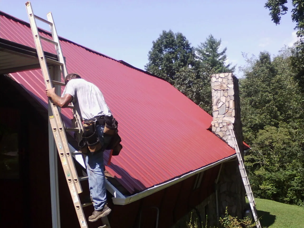Roofer on a ladder beside a red metal roof and stone chimney — Soffit & Fascia Repair by Roofing Brothers in Rockwell City